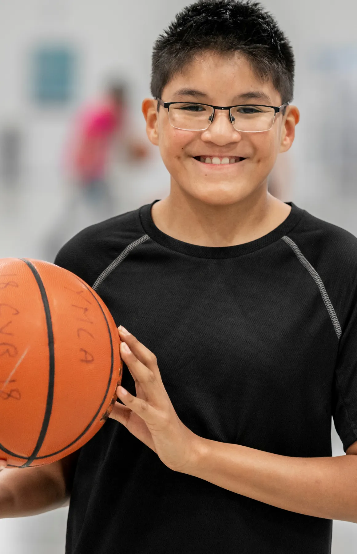 Smiling boy wearing glasses holds a basketball in a gym with a blurred person in the background.