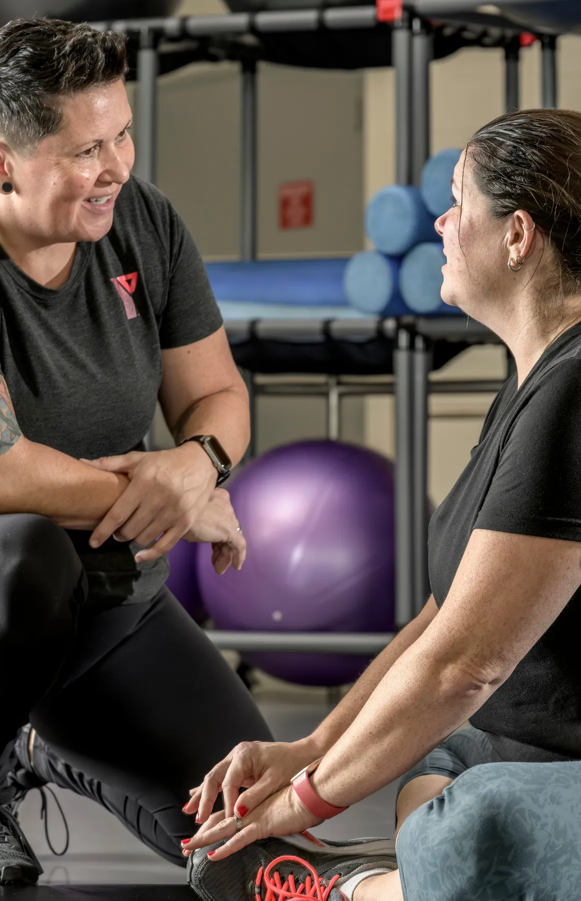 Two women in workout clothes talking in a gym with exercise balls and mats behind them.