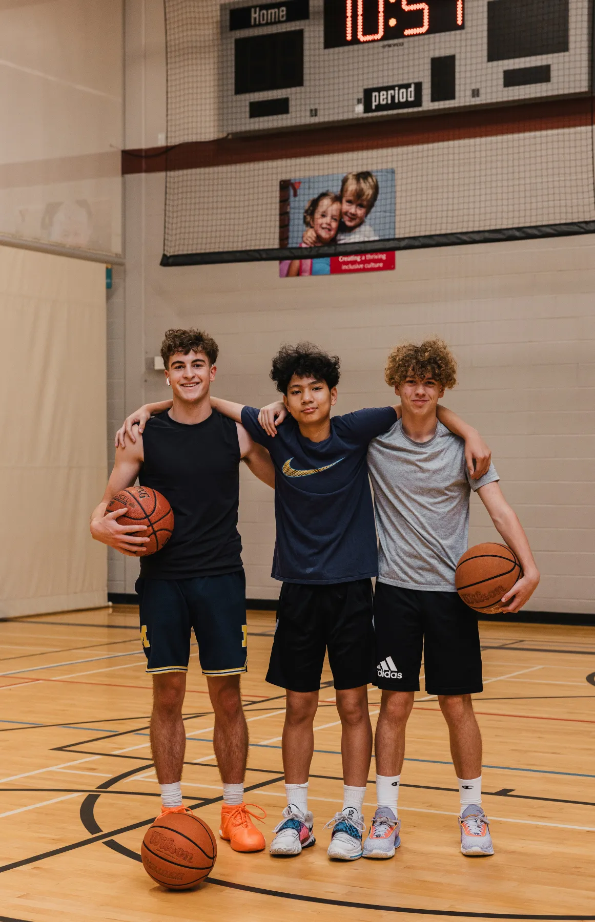 Three teenage boys in a basketball gym, each holding a basketball, smiling with arms around shoulders.