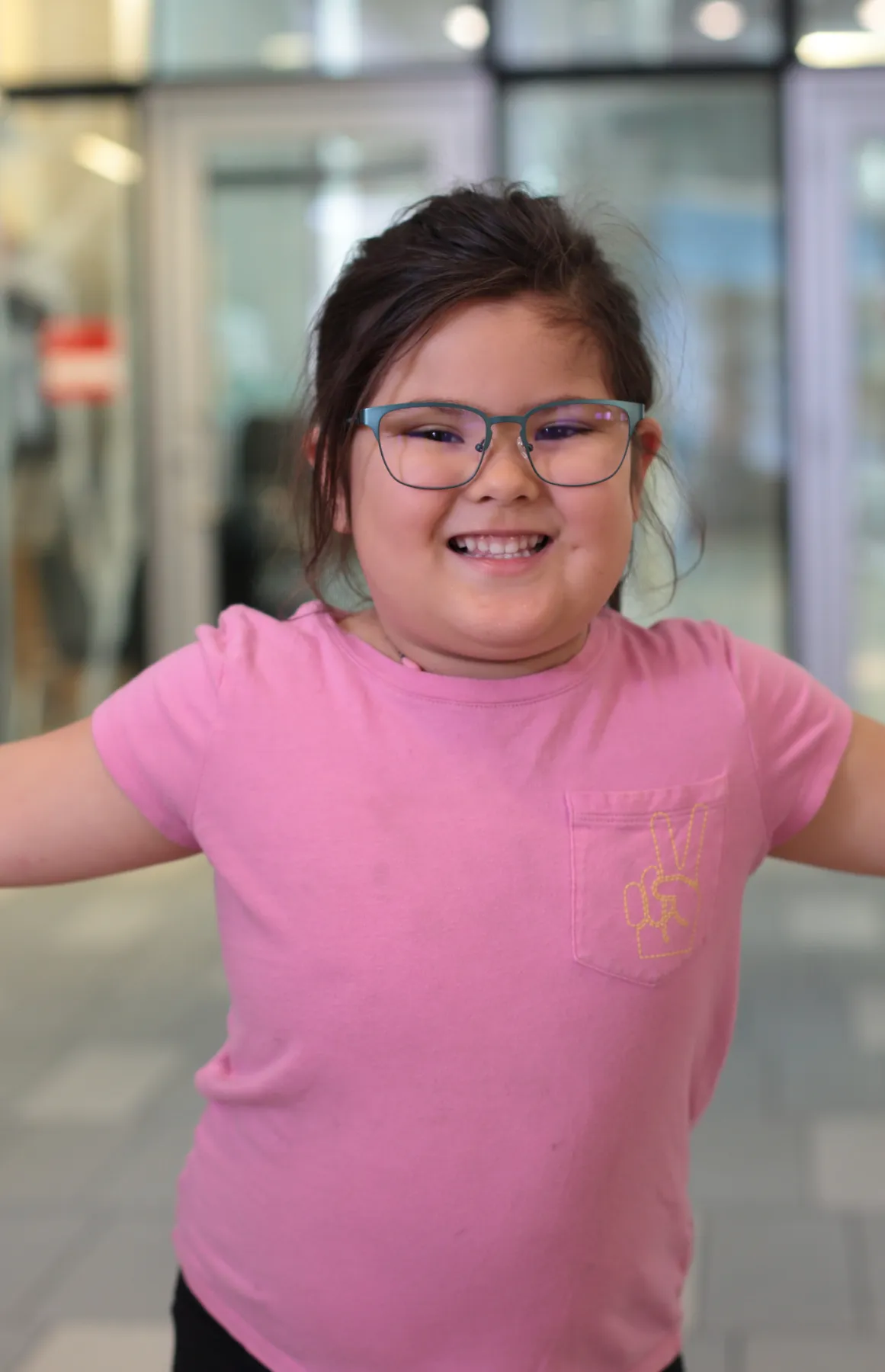 Smiling girl wearing glasses and a pink shirt with a yellow peace sign on the pocket.