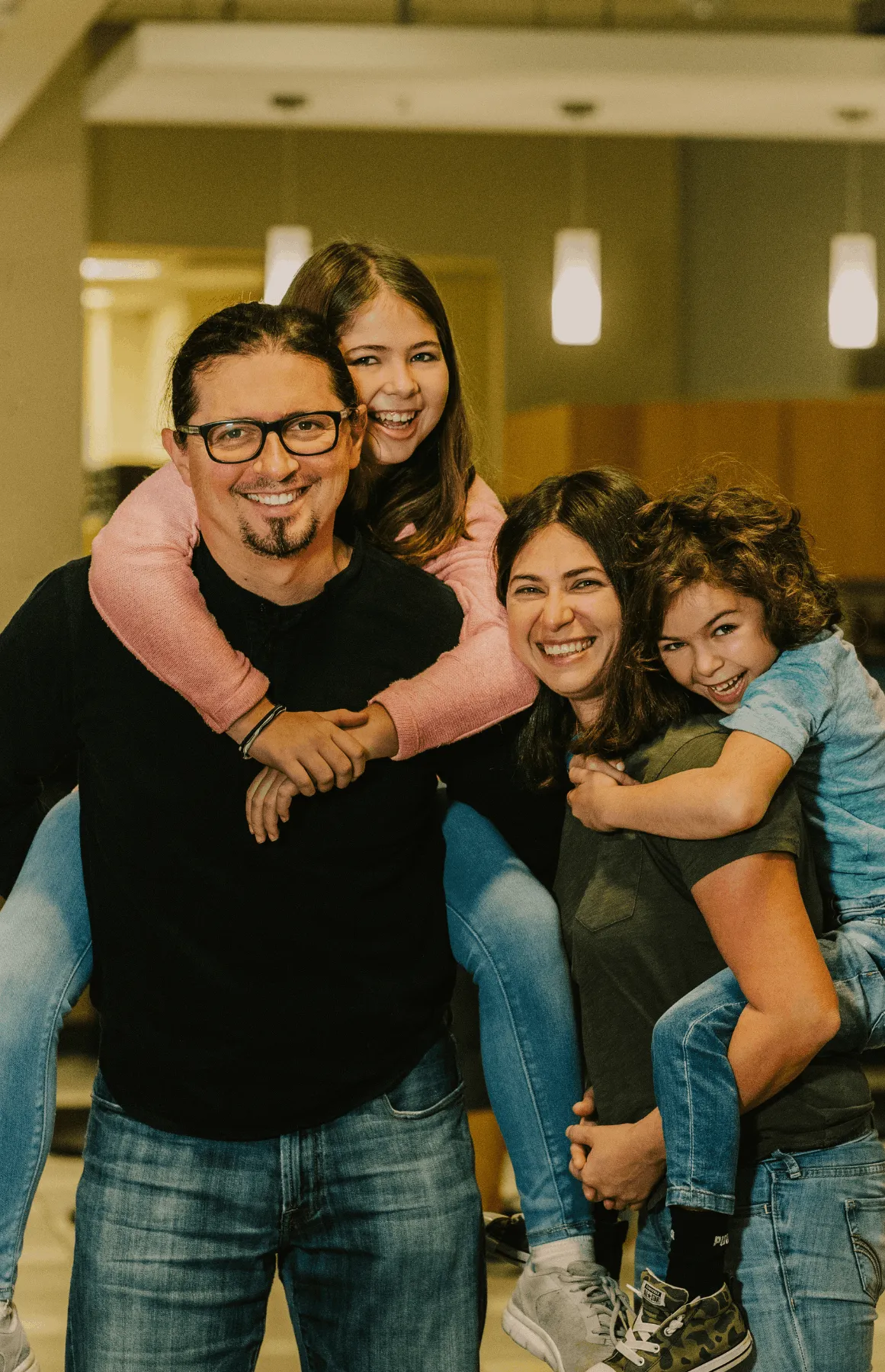 Smiling parents each carrying a child on their back in an indoor setting with warm lighting.