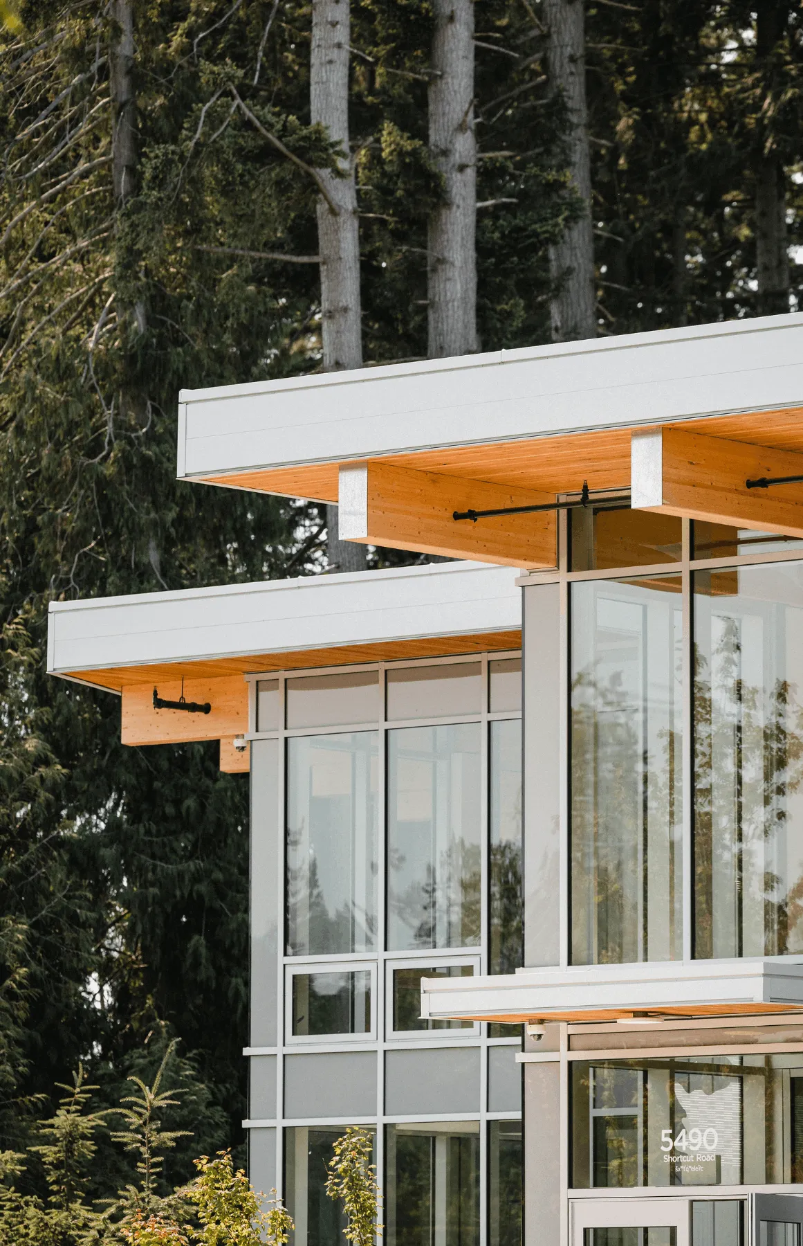 Modern building with large glass windows and wood-paneled overhangs surrounded by tall evergreen trees.
