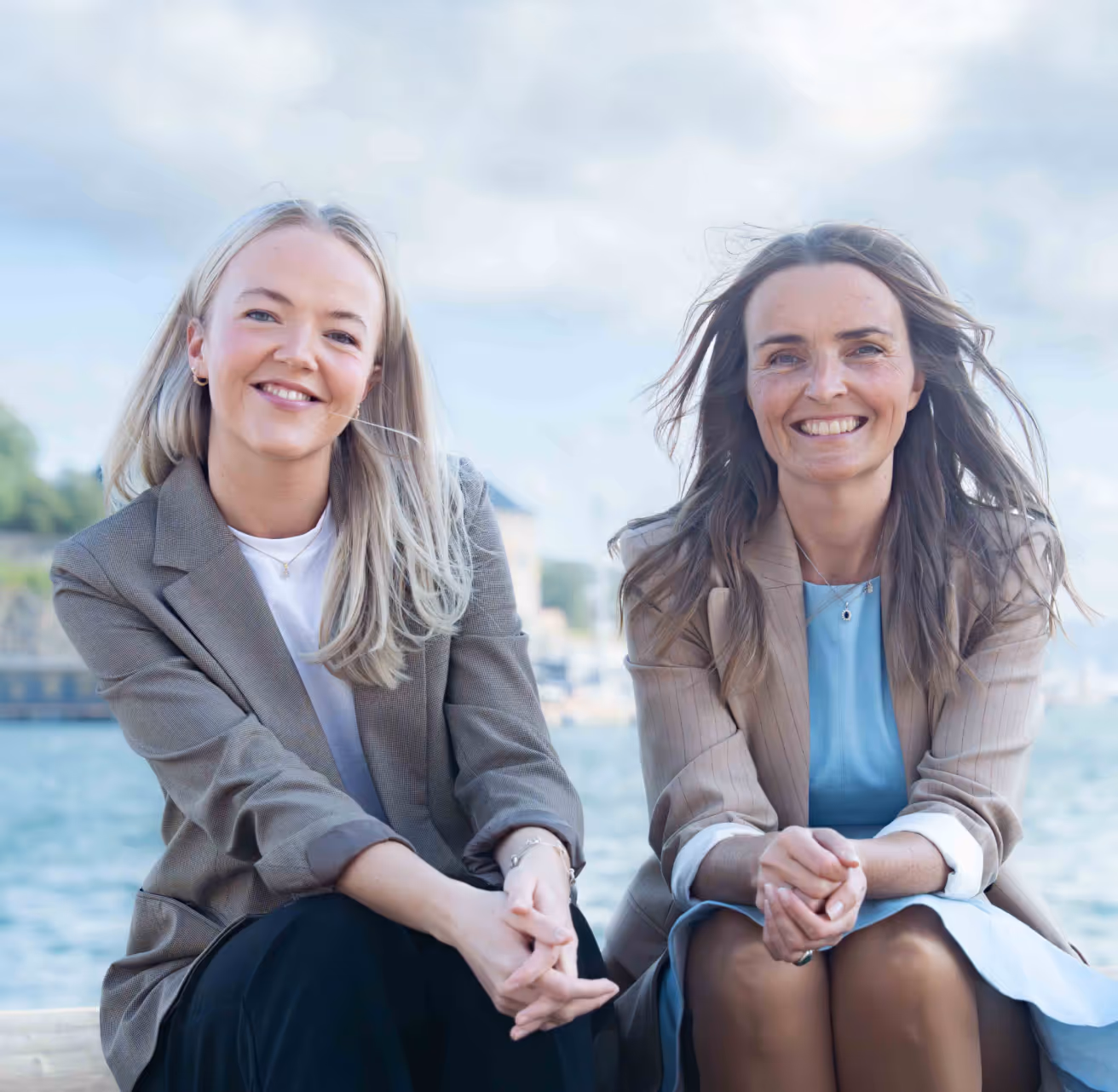 Two smiling women sitting outdoors near water, dressed in blazers with crossed hands.