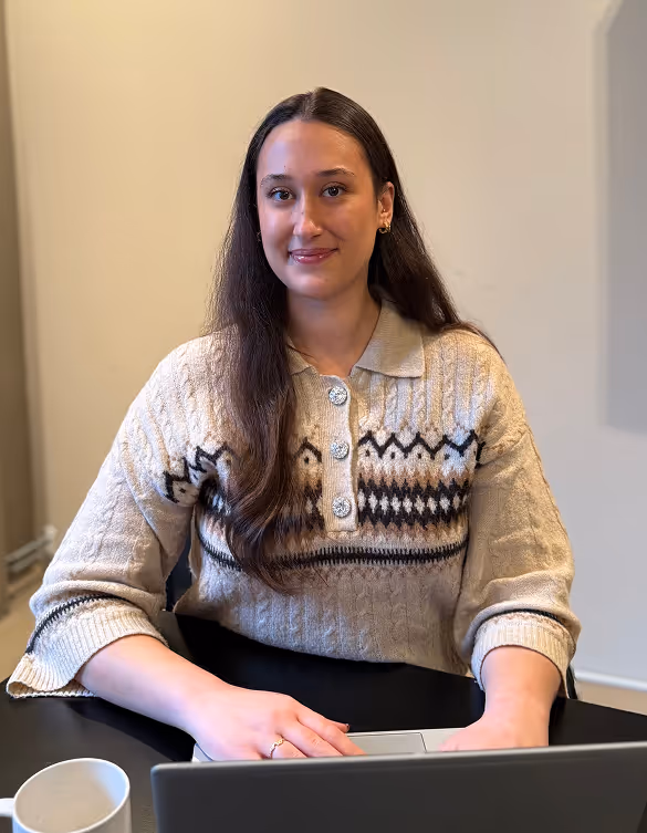 Woman with long brown hair wearing a patterned beige sweater, sitting at a desk with a laptop and a white mug.