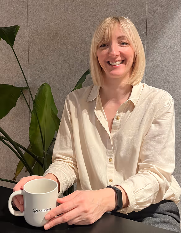 Smiling woman with blonde bob haircut wearing a beige shirt holding a white mug with the 'soliditet' logo, sitting at a table.