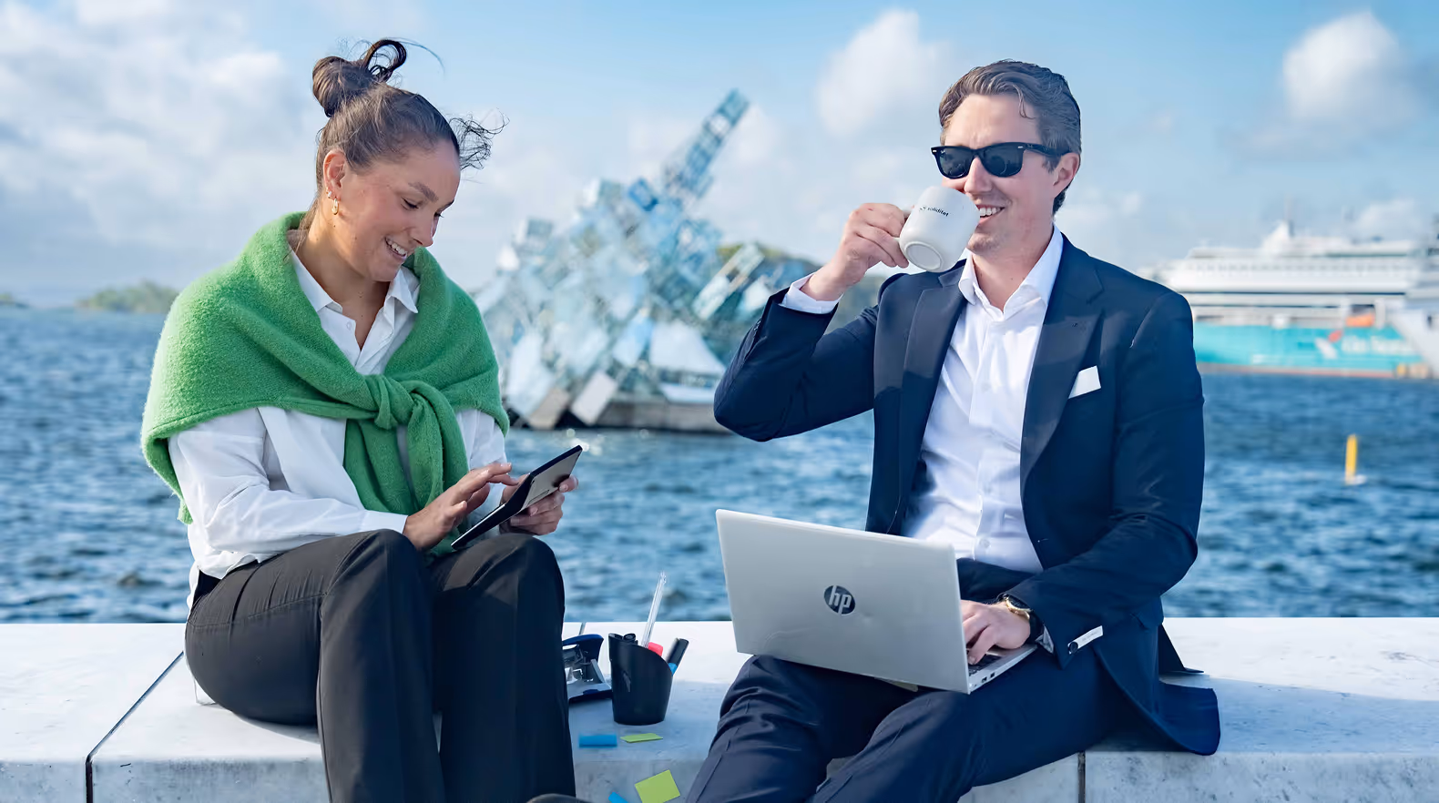 A woman and man sitting outdoors by water, the woman using a tablet while the man works on a laptop and drinks from a mug.