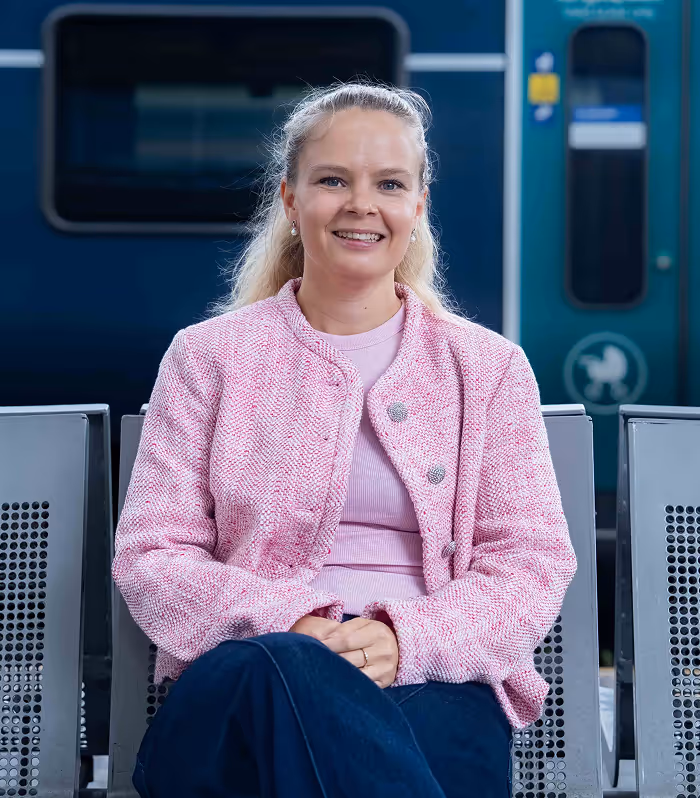 Smiling woman with blonde hair wearing a pink jacket and sitting on a bench at a train station.