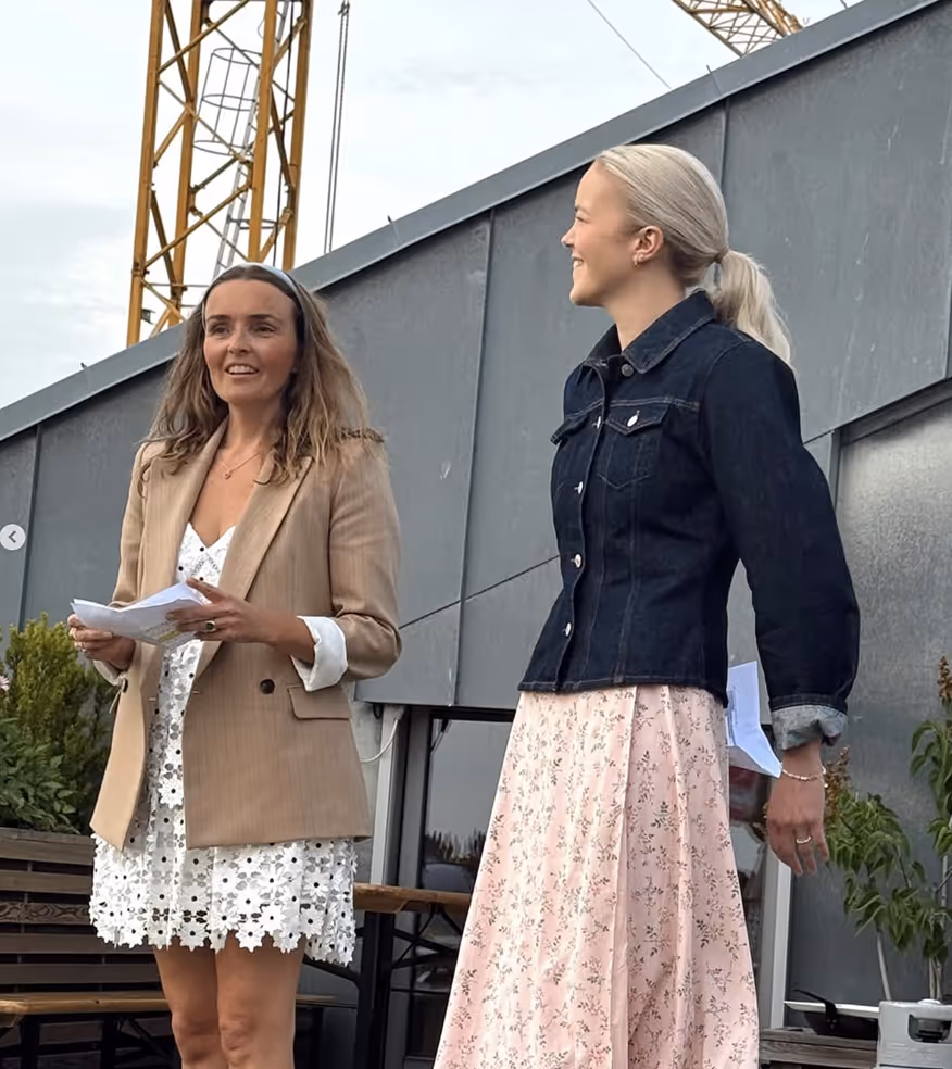Two women standing outdoors; one in a beige blazer and white floral dress holding papers, the other in a dark denim jacket and pink floral skirt smiling and looking to the side.