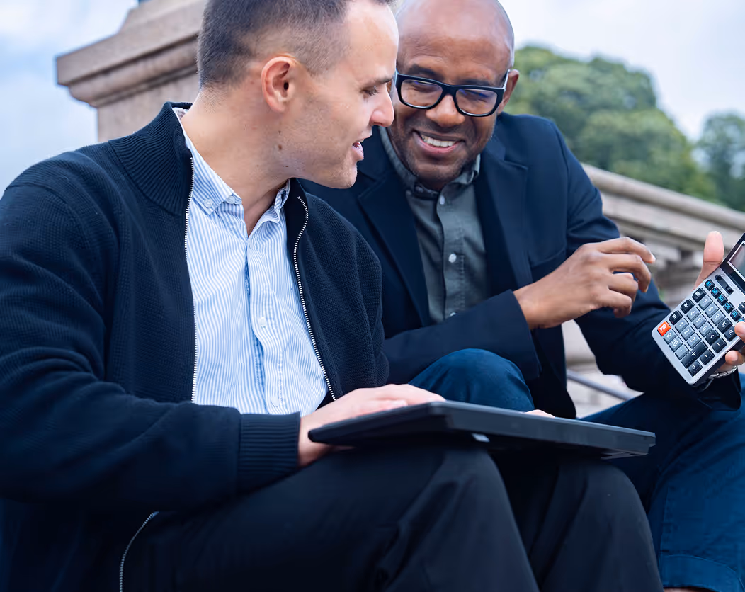 Two men sitting outdoors, one holding a laptop and the other pointing at a calculator while smiling.