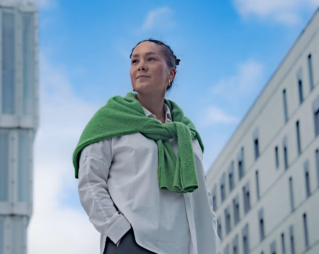 Young woman with a green sweater draped over her shoulders looking confidently to the side against a blue sky background with modern buildings.