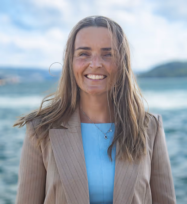 Kvinne med langt blondt hår som smiler foran et vannlandskap med blå himmel.