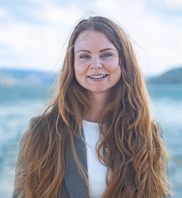 Kvinne med langt, lyst hår og blå øyne smiler foran et vakkert kystlandskap med sjø og fjell i bakgrunnen.