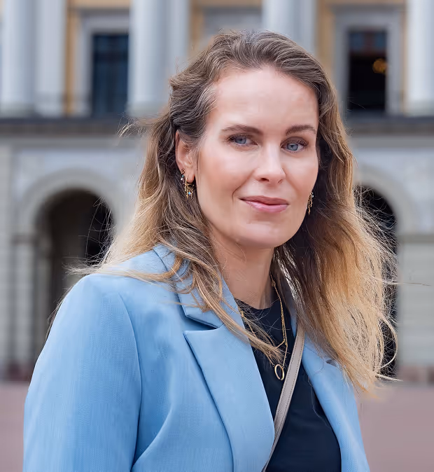 Nærbilde av en kvinne med lyst, bølget hår iført blå jakke utenfor en bygning med buede innganger.