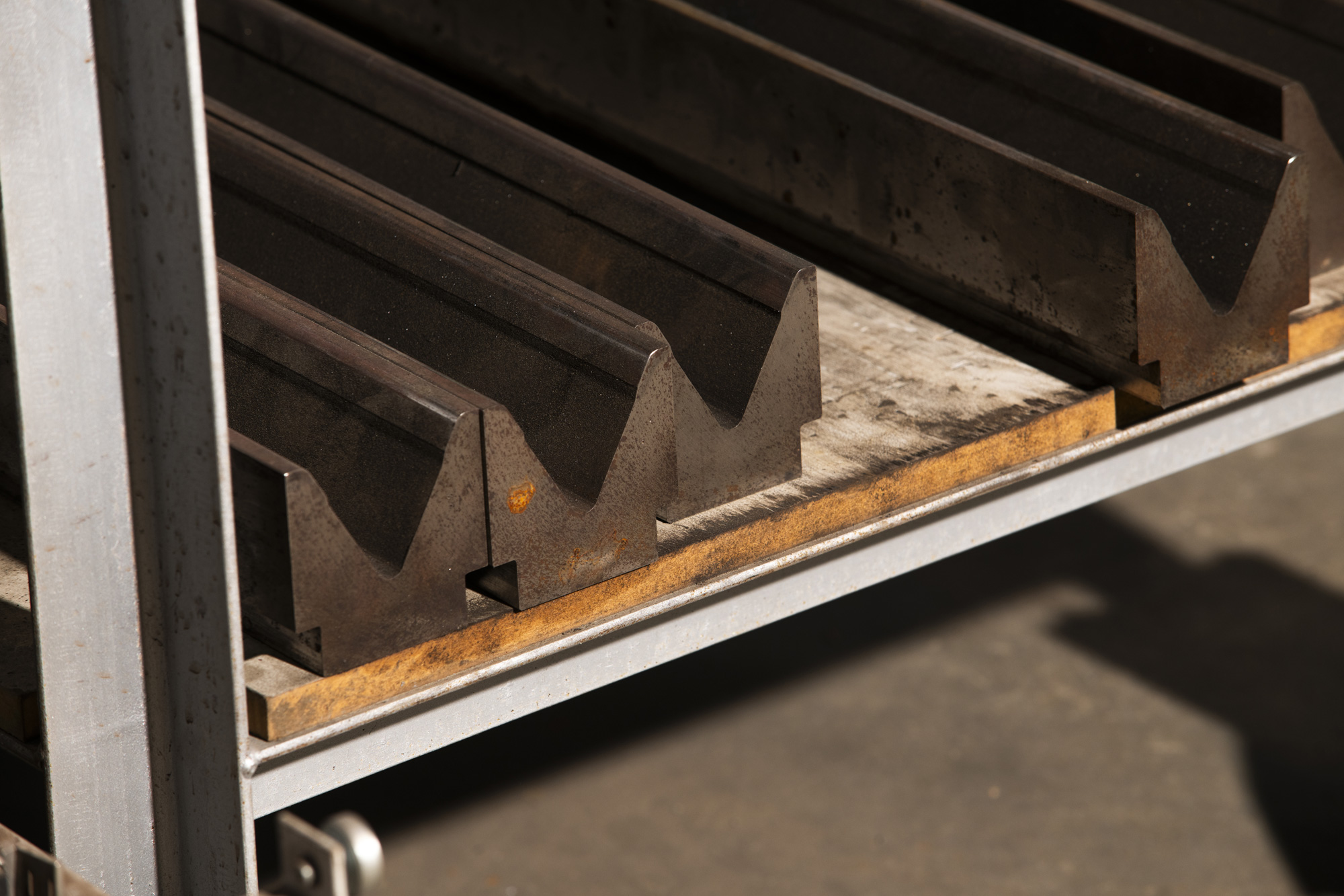 Metal V-shaped press brake dies arranged on a wooden shelf in a metal rack.