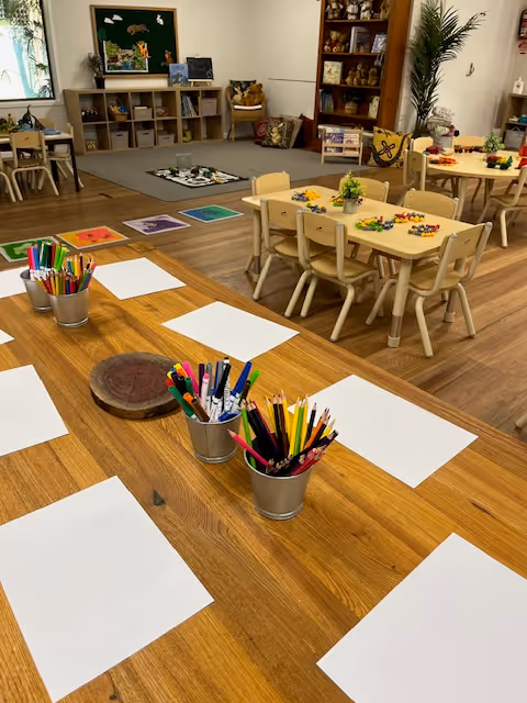 Classroom with tables set up for drawing, featuring paper and buckets of coloured pencils, with additional activity tables and shelves in the background.