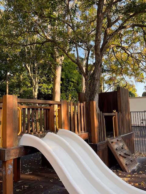 Wooden playground structure with twin white slides set under large leafy trees.