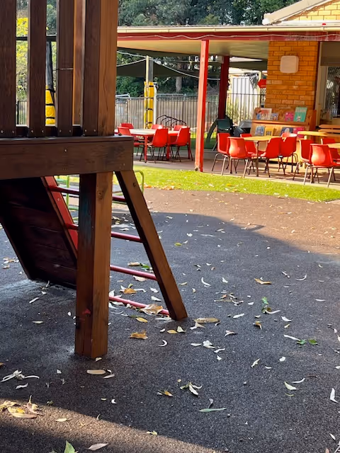 Outdoor play space with part of a wooden climbing structure in the foreground and tables with red chairs under a covered area.