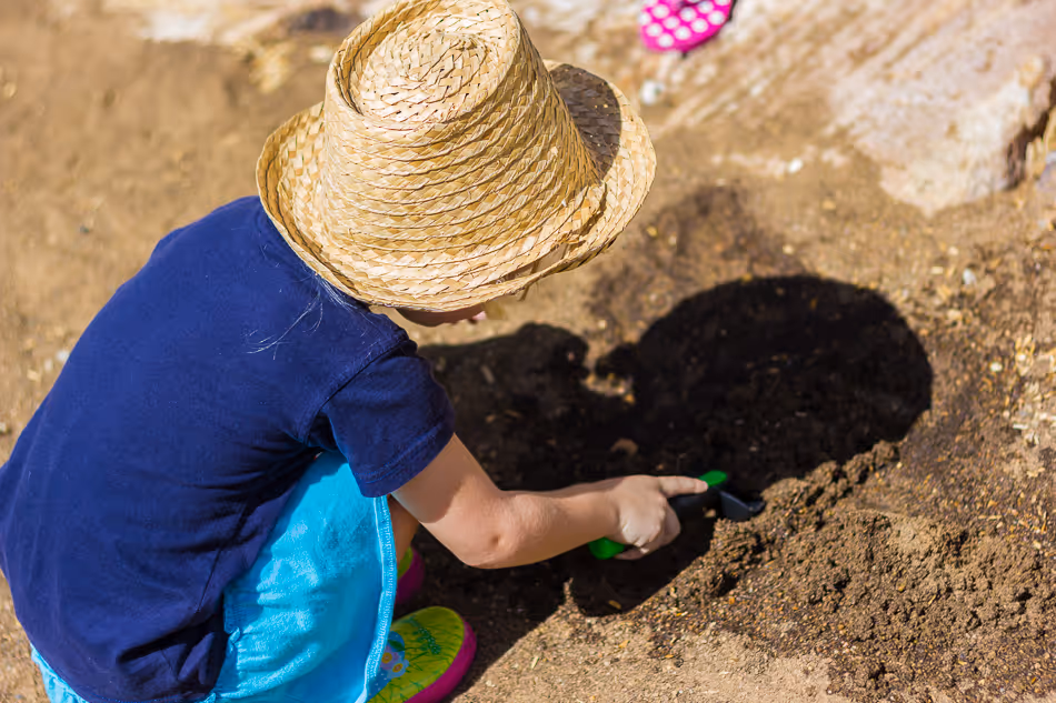Child in a straw hat and blue skirt using a small green trowel to dig in garden soil.