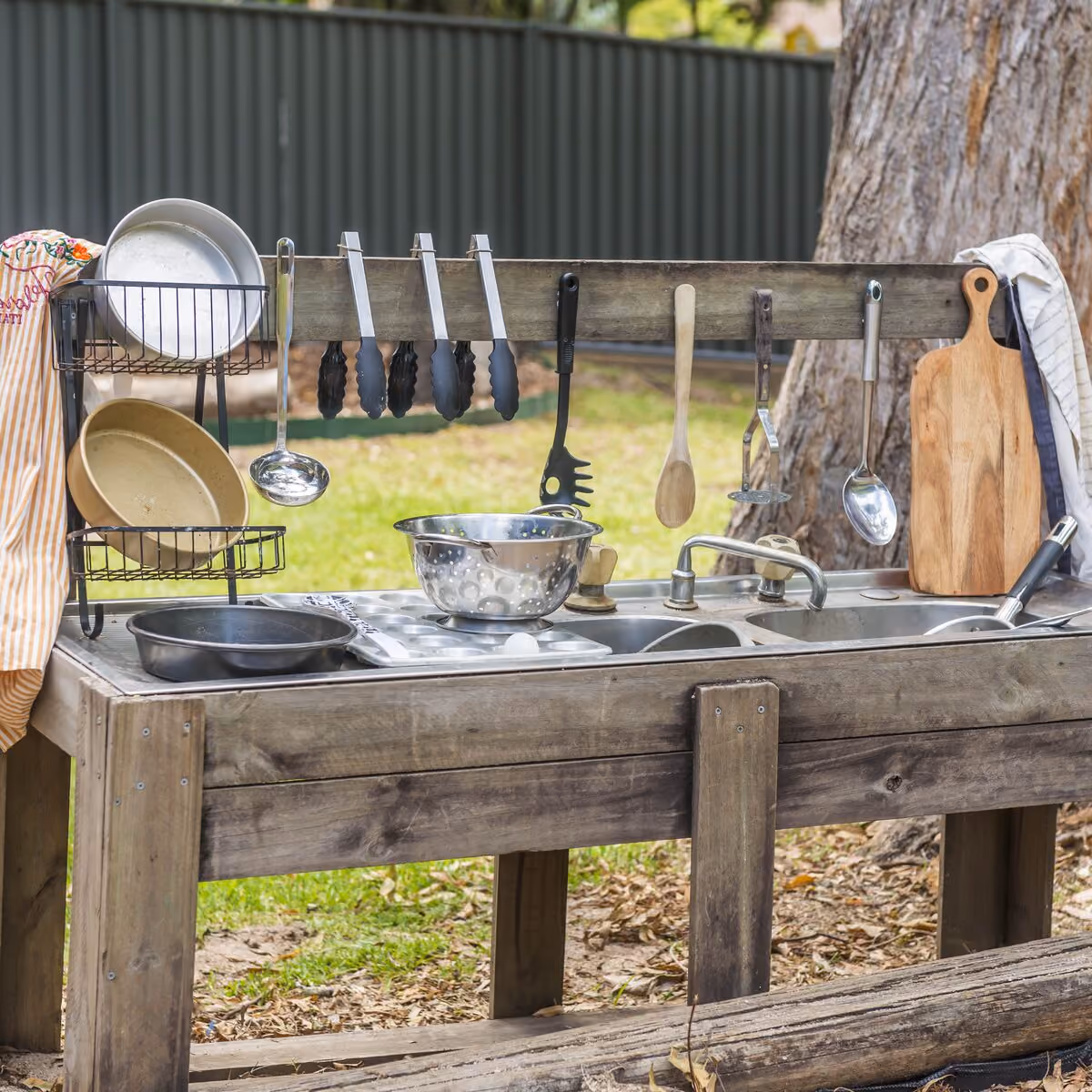 Outdoor mud kitchen with a timber bench, sink, pots, pans and utensils hanging under a large tree.