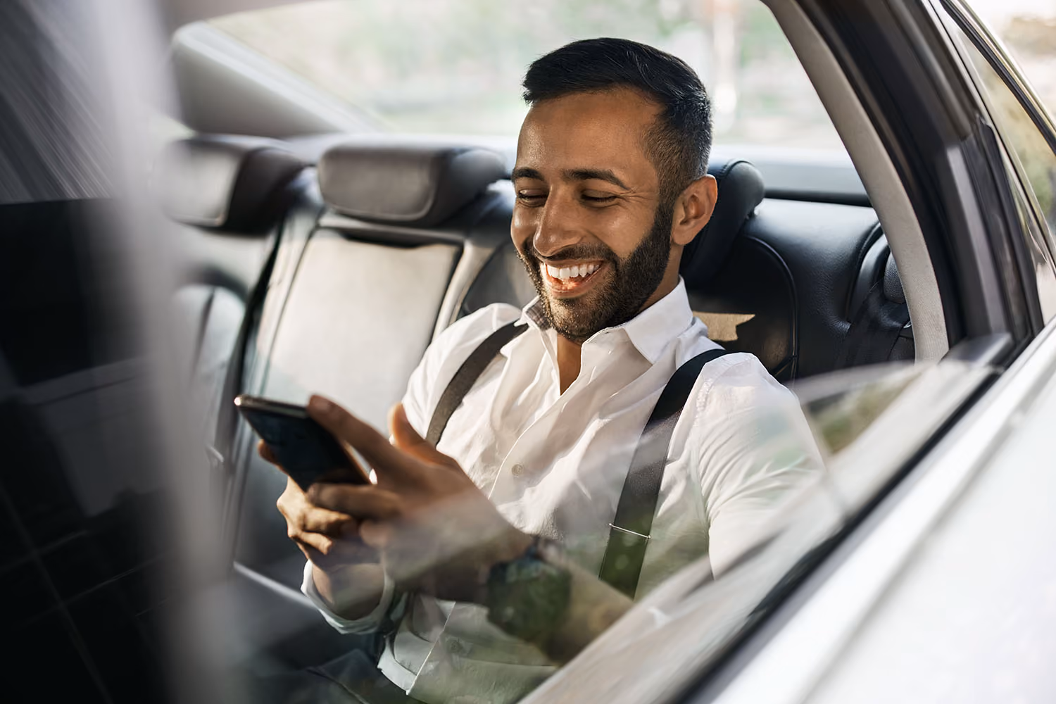 Smiling man in white shirt with suspenders sitting in car backseat looking at his smartphone.