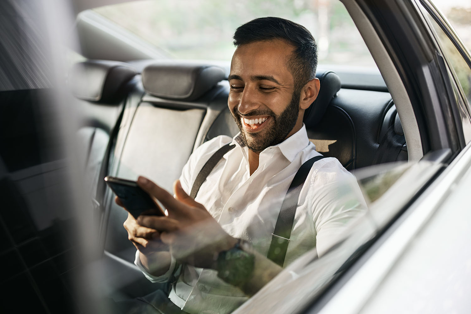 Smiling man in white shirt with suspenders sitting in car backseat looking at his smartphone.