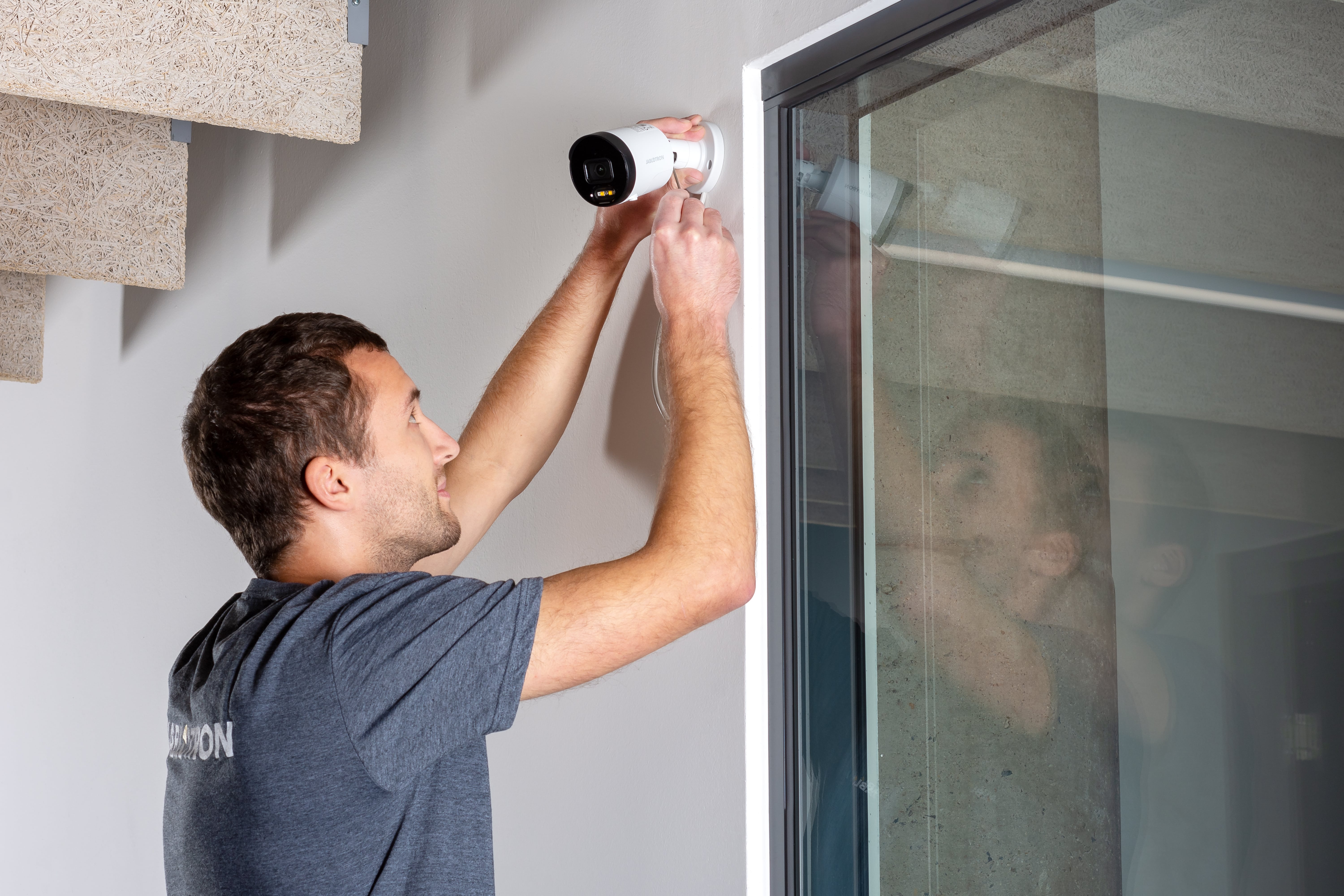Mann installiert eine Sicherheitskamera an einer weißen Wand neben einem Fenster.