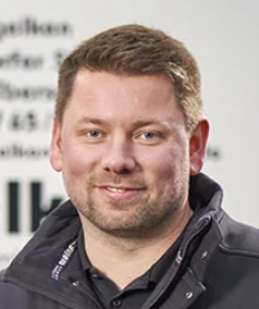 Porträt eines lächelnden Mannes mit kurzen braunen Haaren und dunkelblauer Jacke vor unscharfem Hintergrund mit Text.