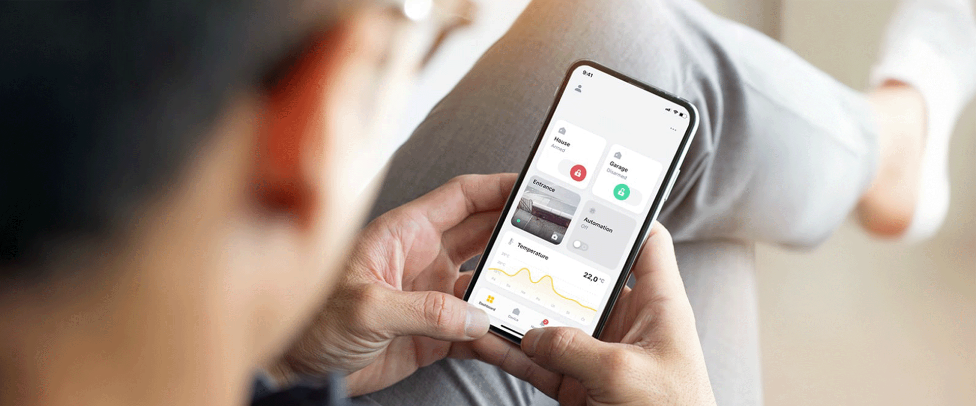 Person hält ein Smartphone mit einer Smart-Home-App, die den Status von Haus und Garage sowie Temperatur und Automatisierung anzeigt.
