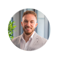 Porträt eines lächelnden Mannes mit kurzem braunem Haar und Bart, der einen hellgrauen Anzug und weißen Hemd trägt.