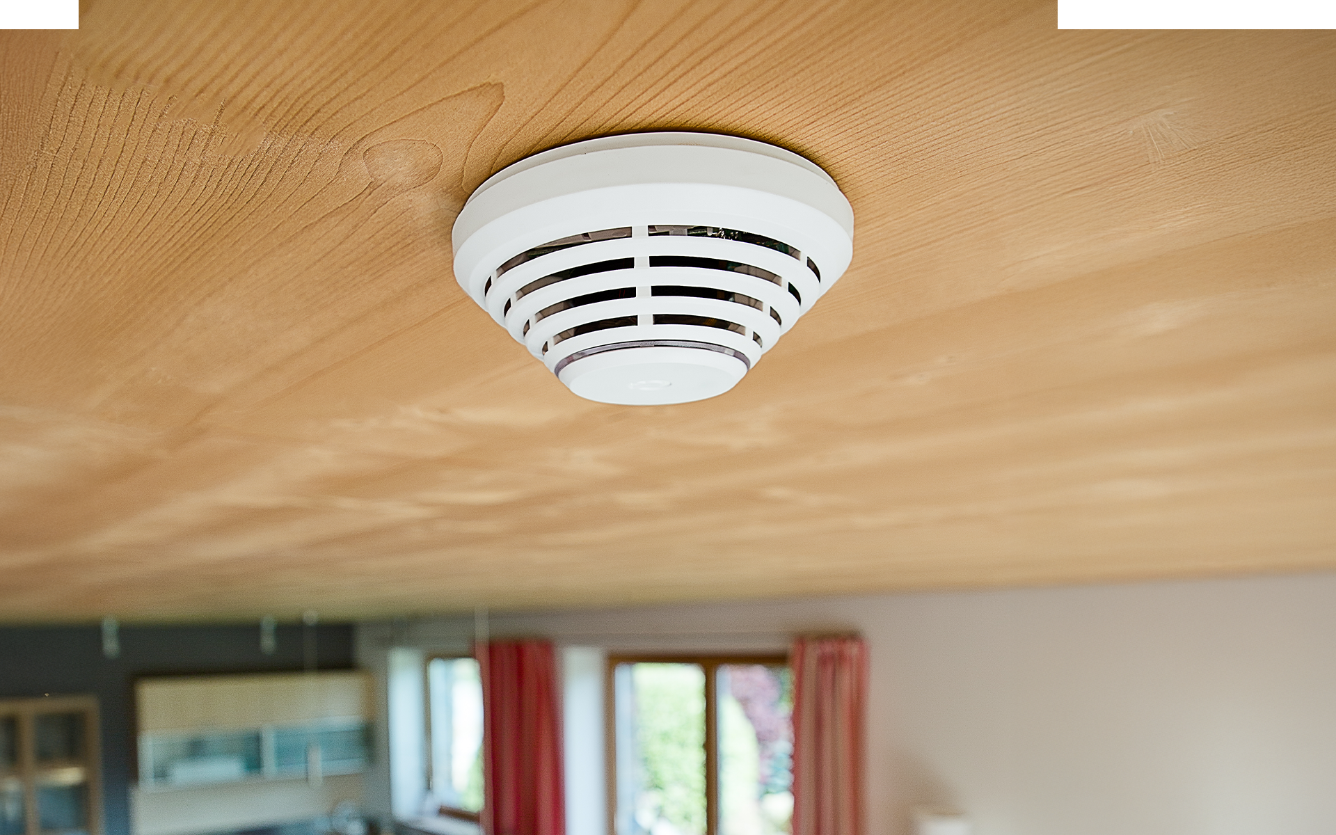 Rauchmelder an einer Holzdecke in einem hellen Raum mit Fenstern und roten Vorhängen.