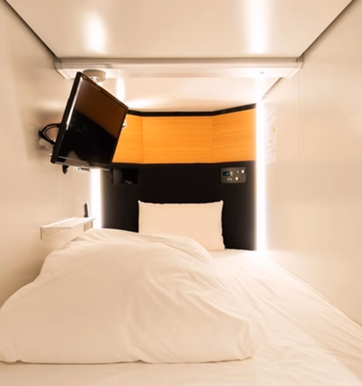 Compact sleeping pod with white bedding, a wall-mounted TV, and ambient lighting around the headboard.