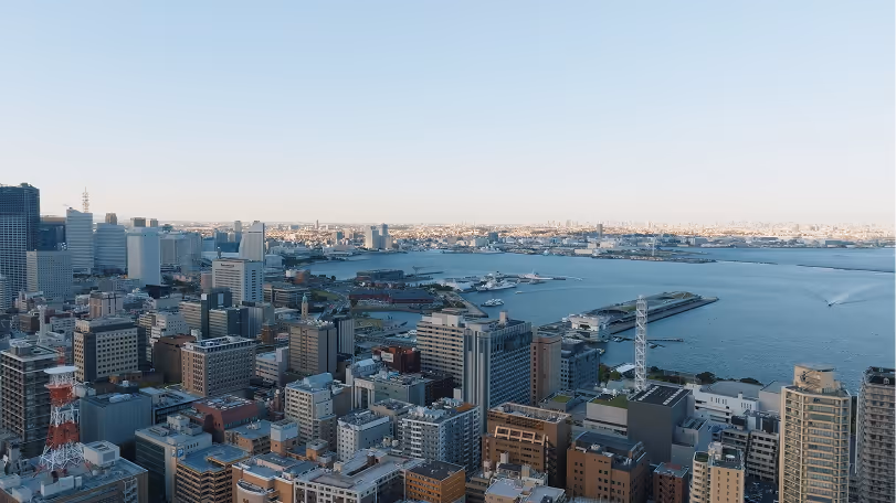 Panoramic cityscape of a waterfront urban area with numerous modern buildings and a pier extending into the water under a clear sky.