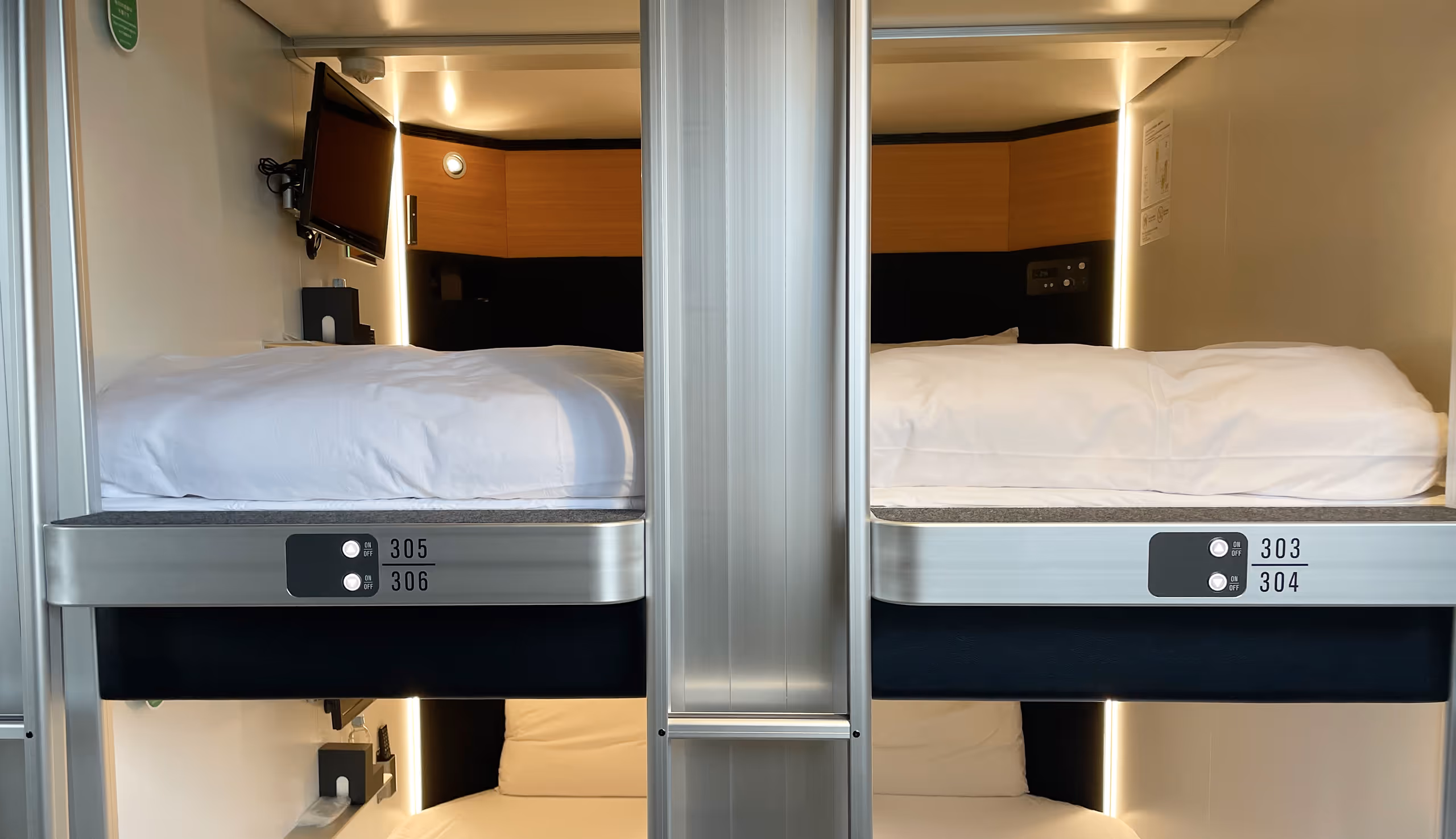 Four neatly made bunk beds in a compact capsule hotel room, each with white bedding, small control panels, and wall-mounted TV.