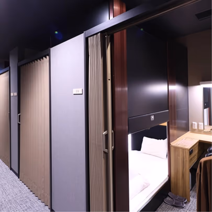 Interior view of a capsule hotel with a small sleeping pod featuring a bed, pillows, desk, slippers, and a curtain entrance.