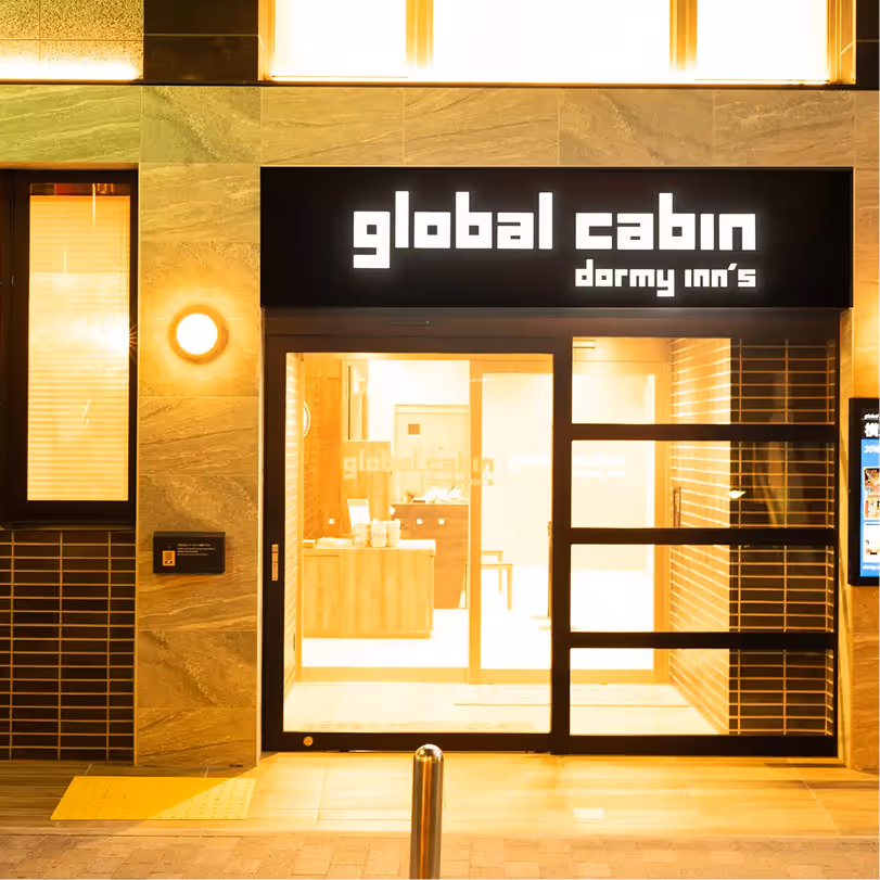 Entrance of global cabin dormy inn's lit warmly at night with a glass sliding door and illuminated sign above.