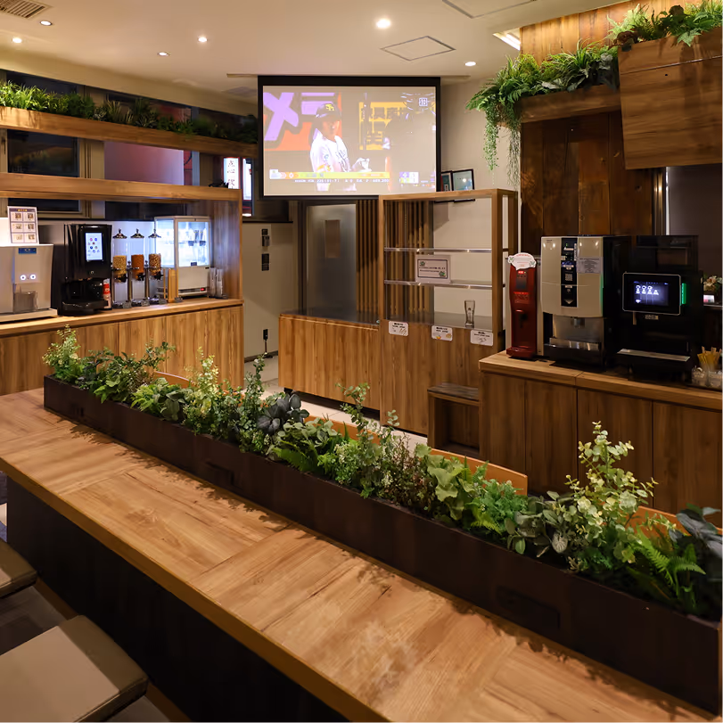 Modern lounge area with wooden counters, a long wooden table with green plants, beverage dispensers, coffee machines, and a large screen showing a sports event.