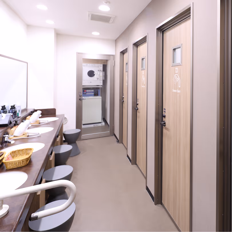 Interior of a clean public shower room with wooden doors labeled 'Shower Room,' round stools, sinks, and a laundry machine at the back.