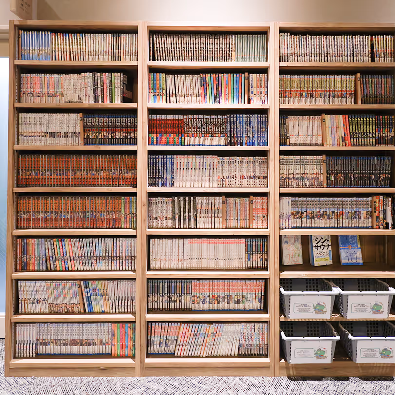 Three wooden bookshelves filled with neatly arranged manga volumes and a few books, with four white storage bins on the bottom right shelf.