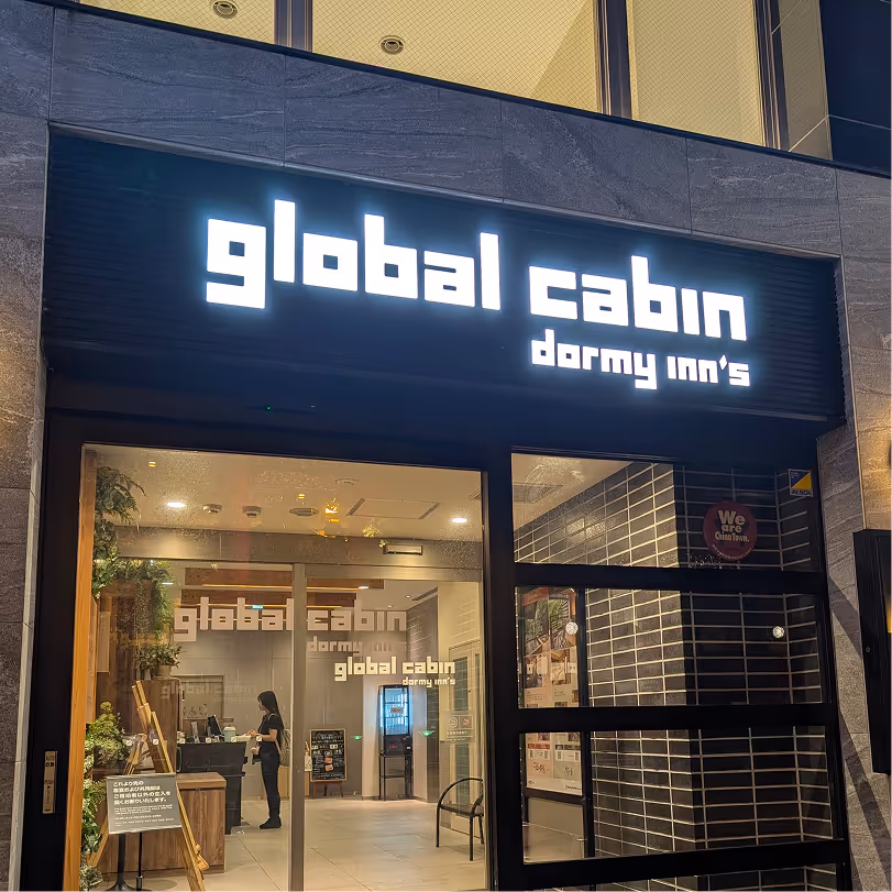 Entrance of global cabin dormy inn's hotel with illuminated sign and glass doors showing the reception area inside.