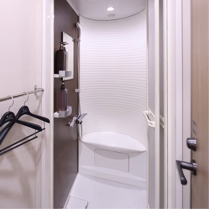 Compact white shower interior with curved wall, built-in seat, two mounted soap dispensers, and black hangers on a towel rack outside.