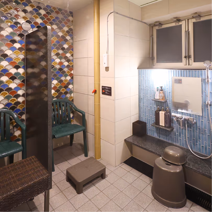 Indoor bathing area with colorful tiled wall, plastic chairs, showerhead, mirror, and bath stools.