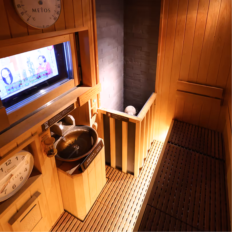 Interior of a wooden sauna with benches, a clock, a water basin, and a small window showing a TV screen.