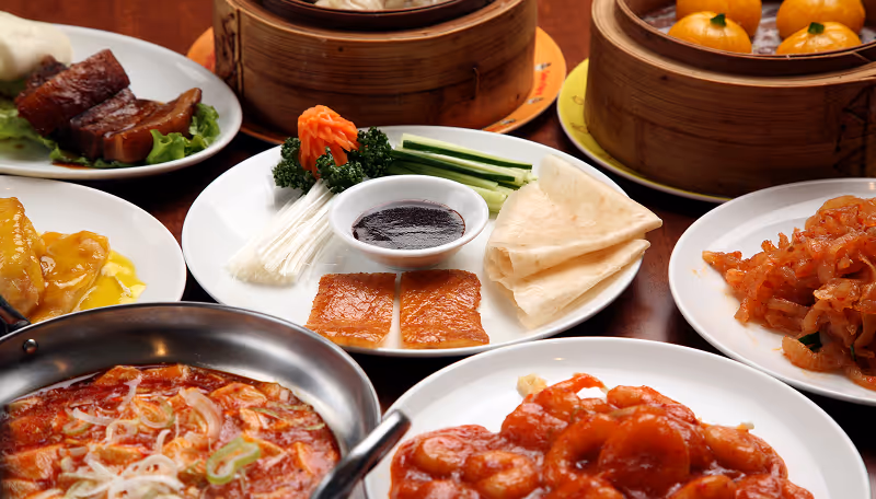 Variety of Chinese dishes including dim sum, glazed meats, prawns in sauce, and vegetables arranged on white plates around a wooden table.