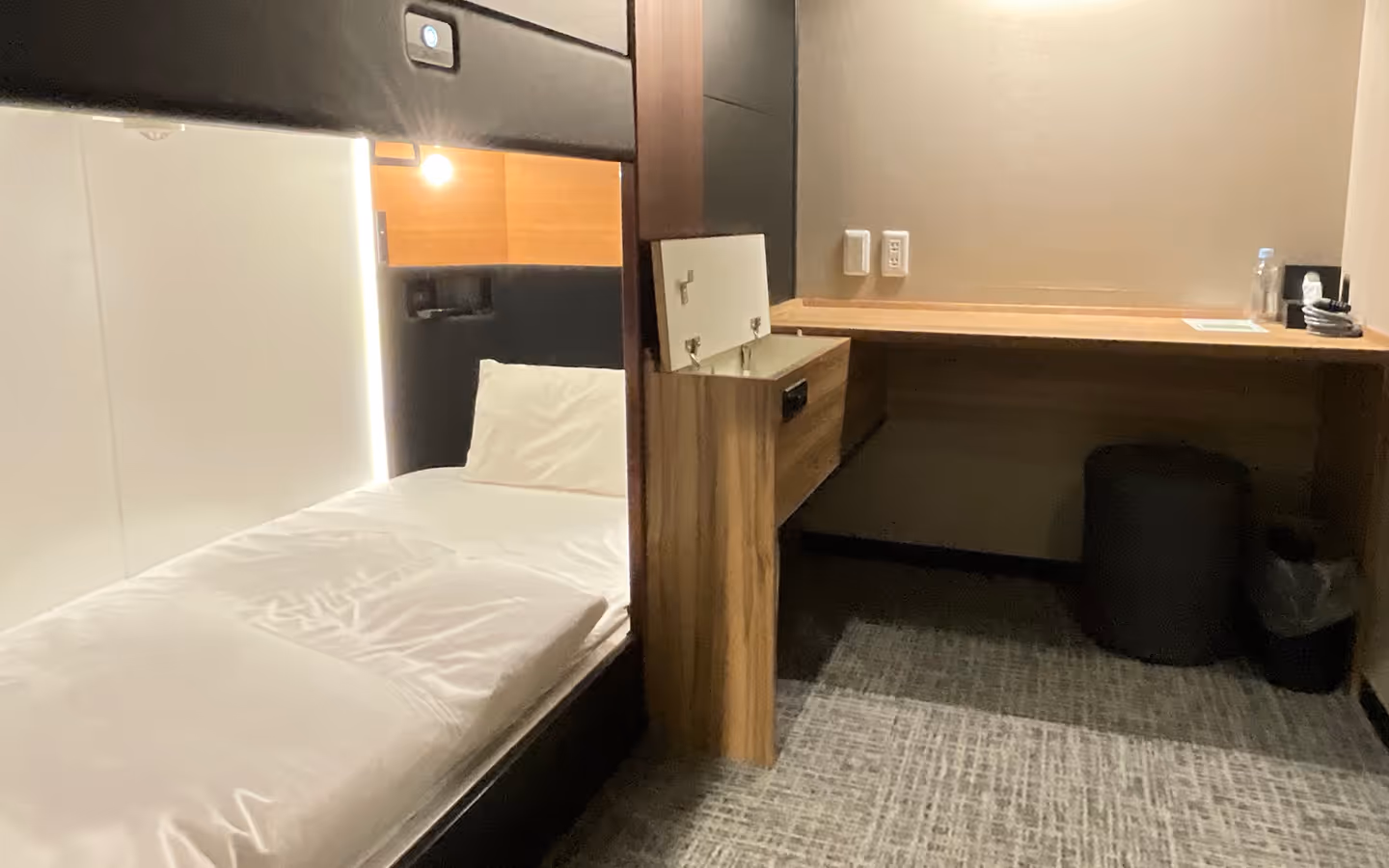 Compact hotel room with a single bed, white bedding, a small desk, a black stool, and a wall lamp.