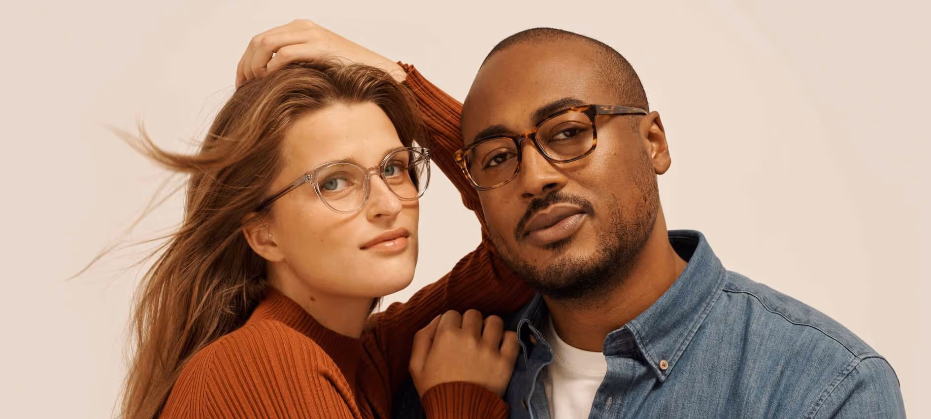 Portrait of a woman in an orange sweater and clear glasses leaning on a man wearing a denim shirt and tortoiseshell glasses.