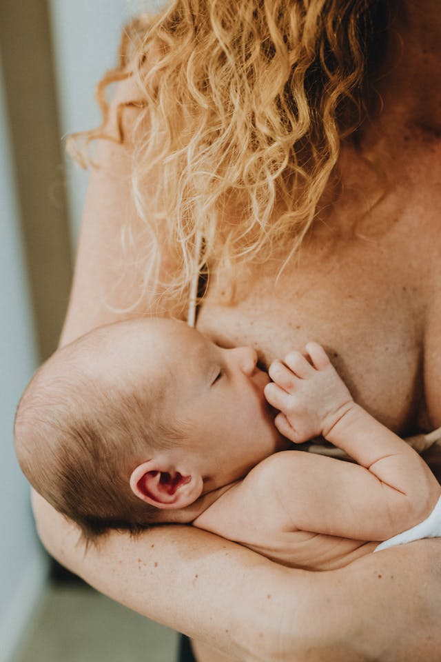 Newborn baby breastfeeding while being held by a person with curly blonde hair.