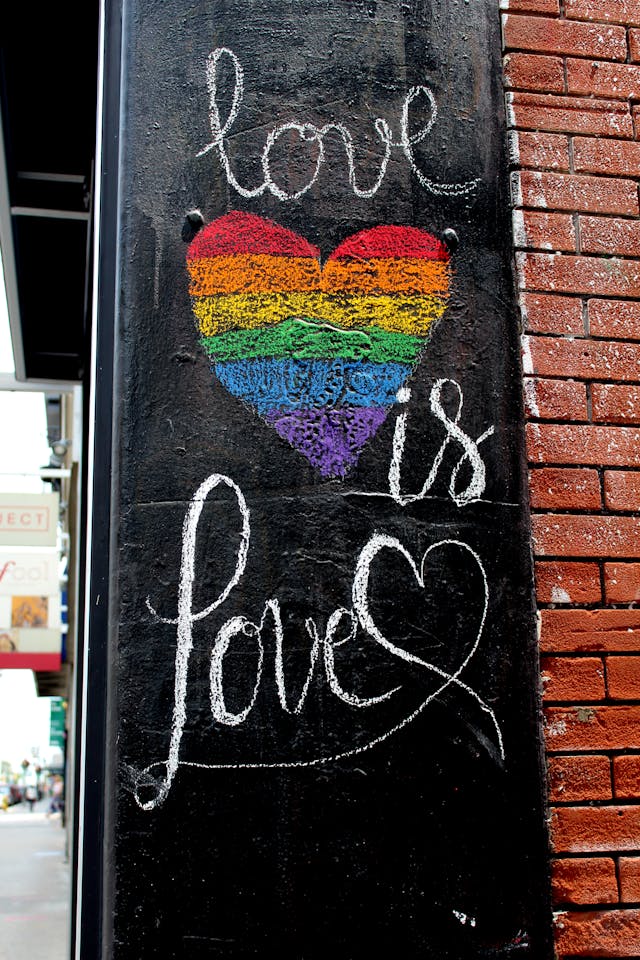 Street art with the phrase 'love is love' and a heart painted in rainbow colors on a black wall next to a red brick wall.