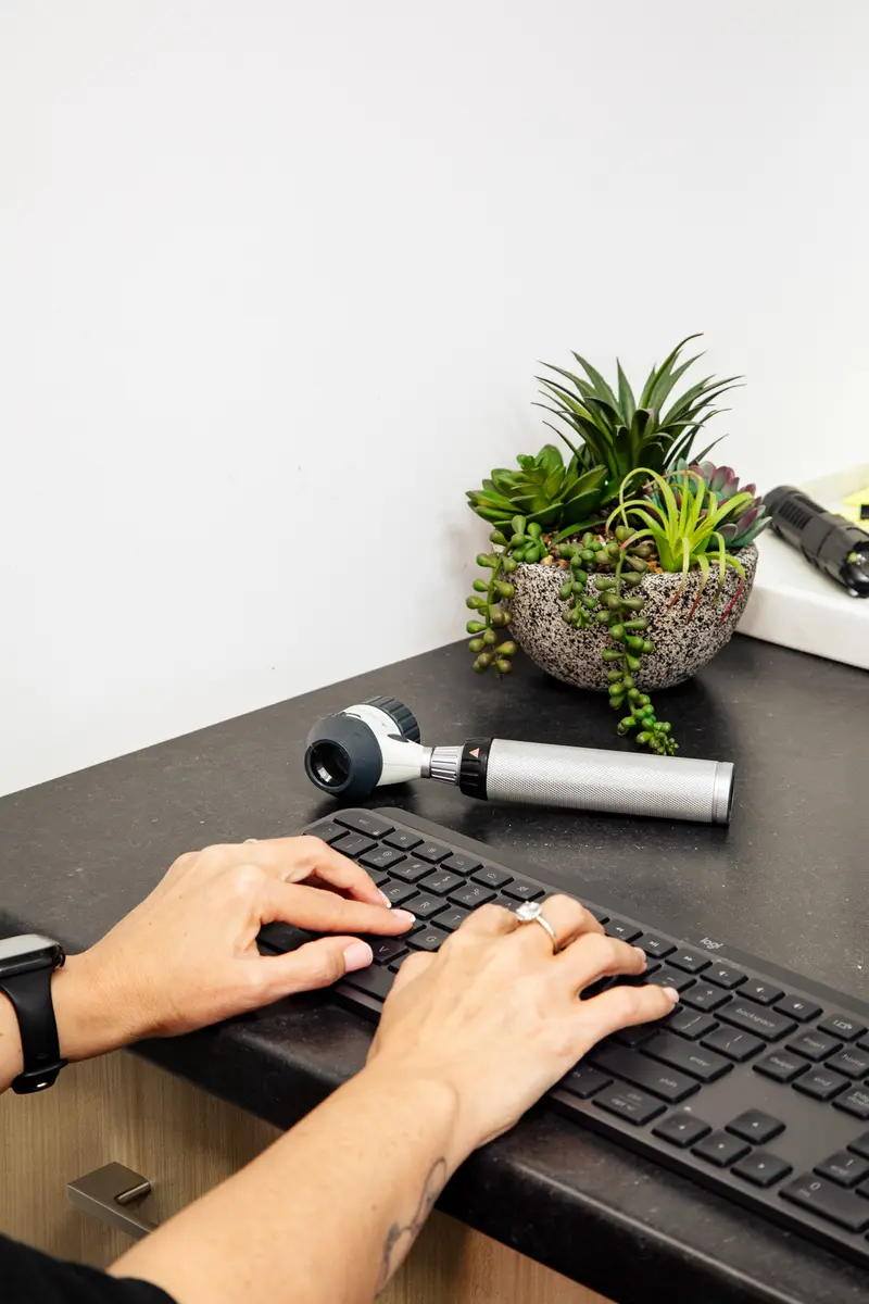 Hands typing on a black keyboard on a desk with a small potted succulent plant and a handheld medical device nearby.