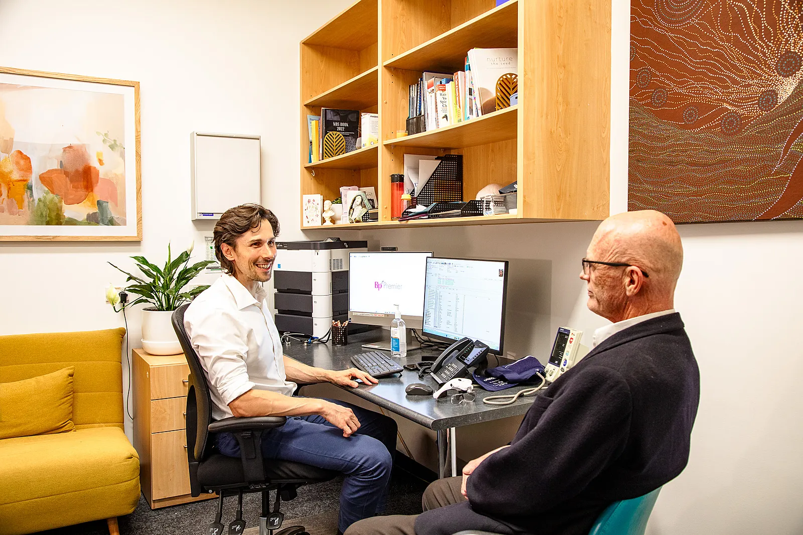 A doctor in a white shirt sits at a desk talking to an elderly male patient in an office with wooden shelves and computer monitors.