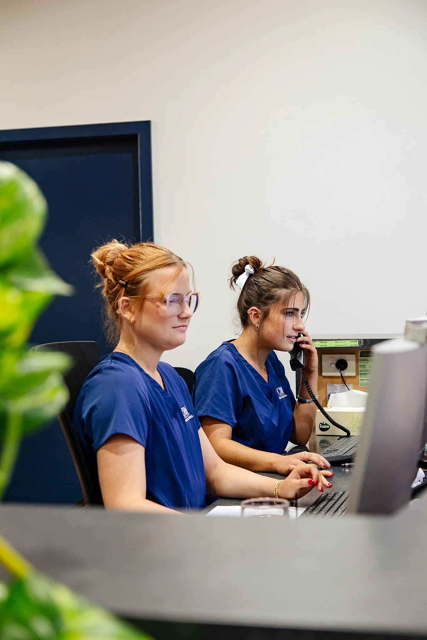 Two women in blue medical scrubs working at a desk with computers, one talking on the phone and the other typing.